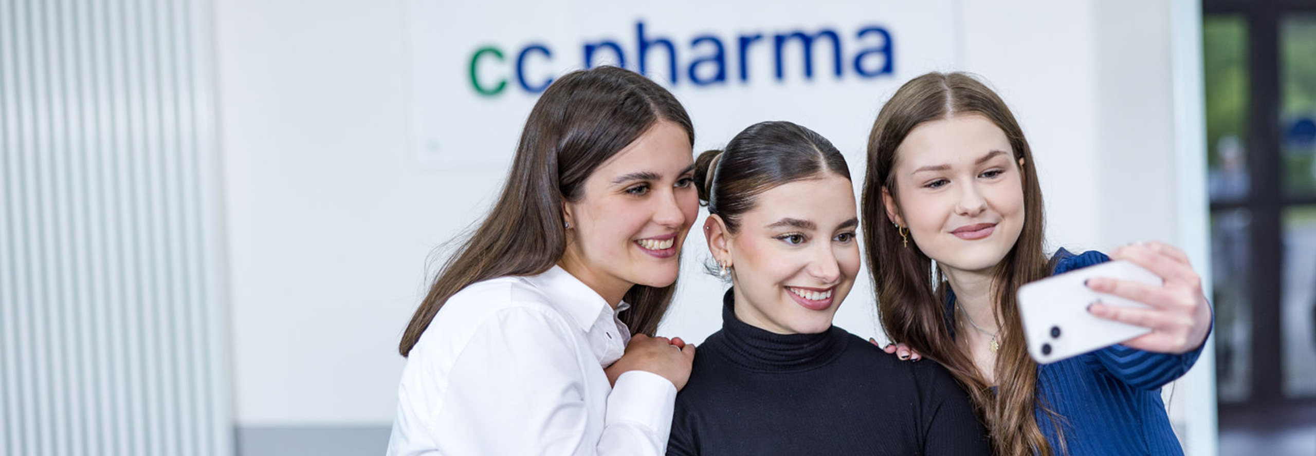 Drei junge Frauen nehmen gemeinsam ein Selfie vor einem Firmenschild von CC Pharma auf. Die Frauen lächeln in die Kamera des Smartphones, das von der Frau rechts gehalten wird.