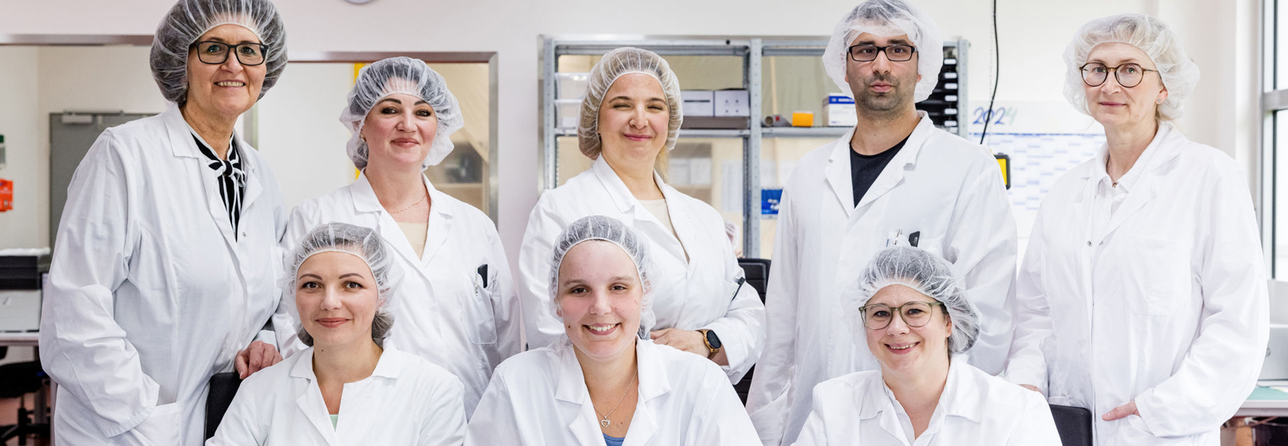 Group photo of eight professionals in a laboratory environment. The seven women and one man are all wearing white lab coats and hairnets. They stand next to one another and are smiling for the camera.