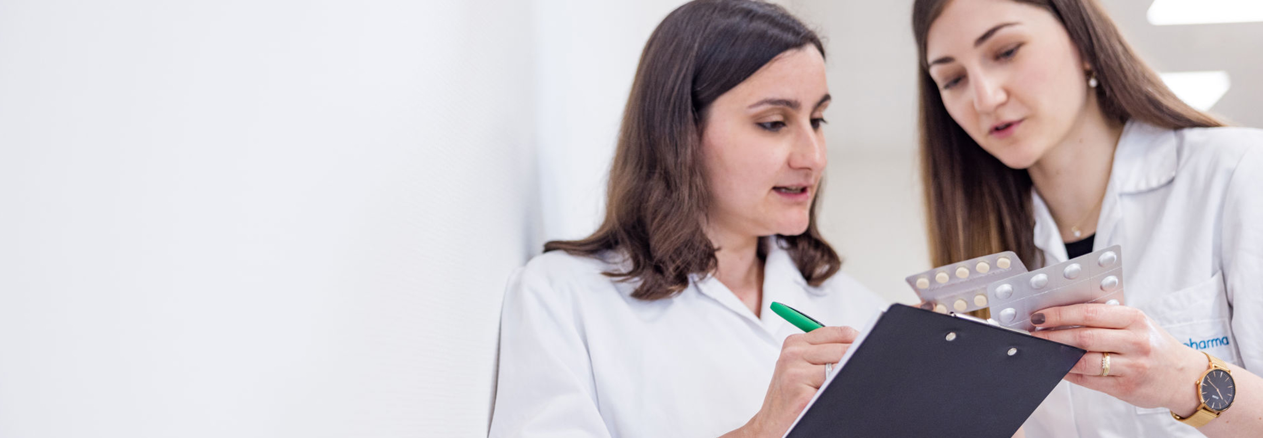 Two women in white coats talk in front of a wall. One woman points to a blister pack while the other makes notes on a clipboard.