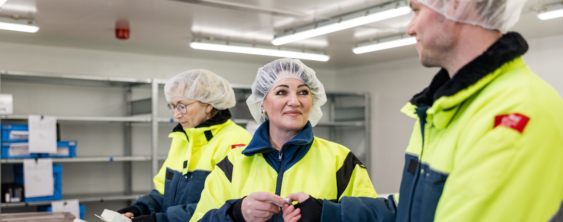 Three people, two women and one man, wearing fluorescent yellow safety jackets and hairnets, are seen in a cold room. The focus is on the woman and man in the foreground who are talking and handing over a piece of paper. The women in the background is looking at a package of medicines.