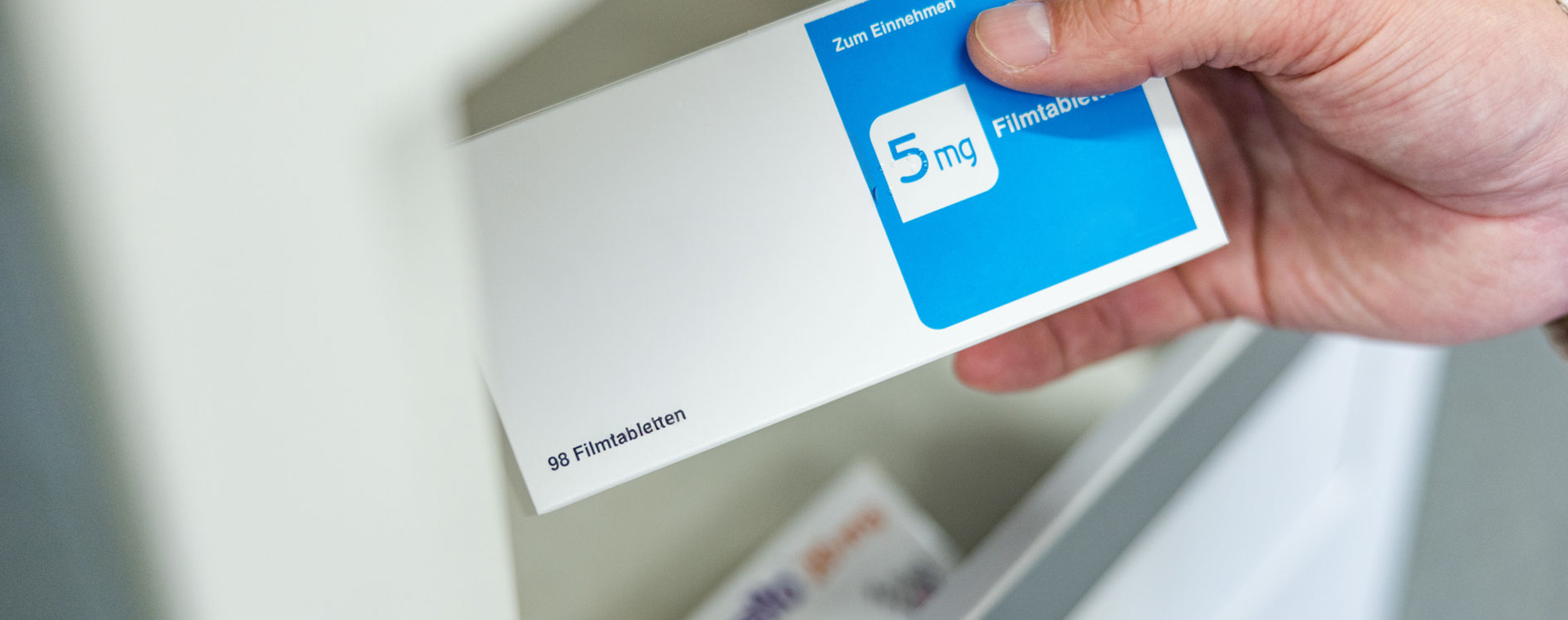 Eine Hand hält eine Medikamentenverpackung mit Filmtabletten. Deutlich erkennbar durch das Etikett mit „5mg“ in blau und weiß. Der Fokus liegt auf der Packung.