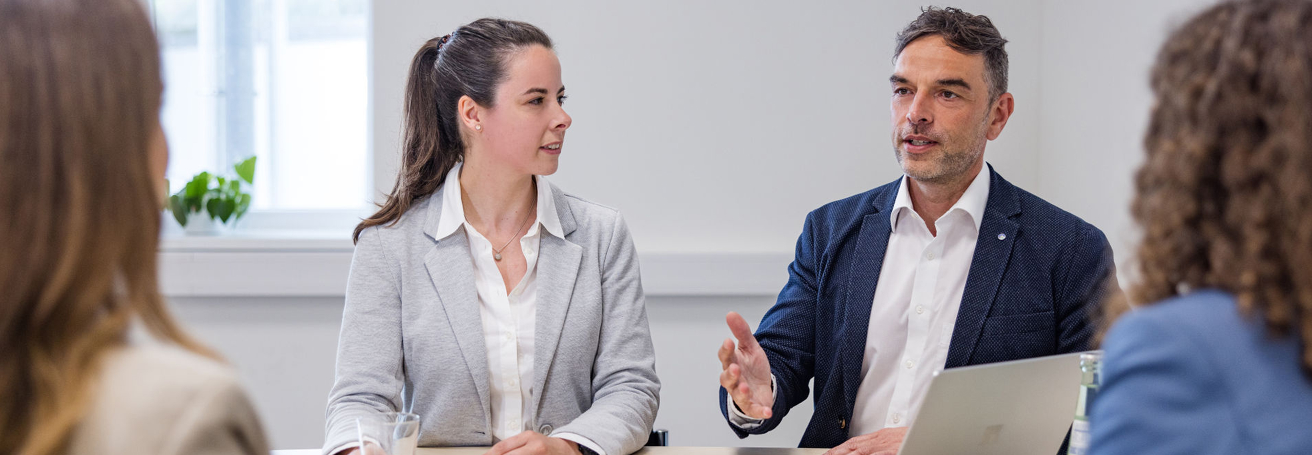 Besprechungssituation in einem hellen Büro, bei der ein Mann und eine junge Frau in Business-Kleidung an einem Tisch sitzen. Der Mann sitzt vor einem Laptopbildschirm, während die Frau ihn anschaut. Am rechten und linken äußeren Rand des Bildes sind zwei weitere Personen von hinten zu erkennen