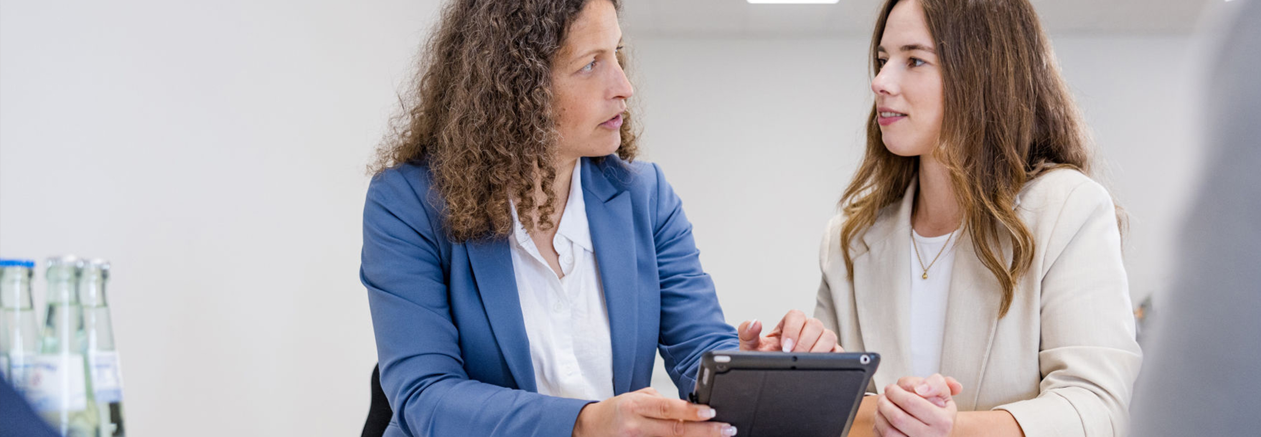 Zwei professionell gekleidete Frauen sitzen an einem Tisch und schauen einander an. Die linke Frau trägt einen blauen Blazer, hält ein Tablet in der Hand und zeigt darauf, während die andere Frau in einem beigen Blazer ihre Hände vor sich auf dem Tisch liegen hat.