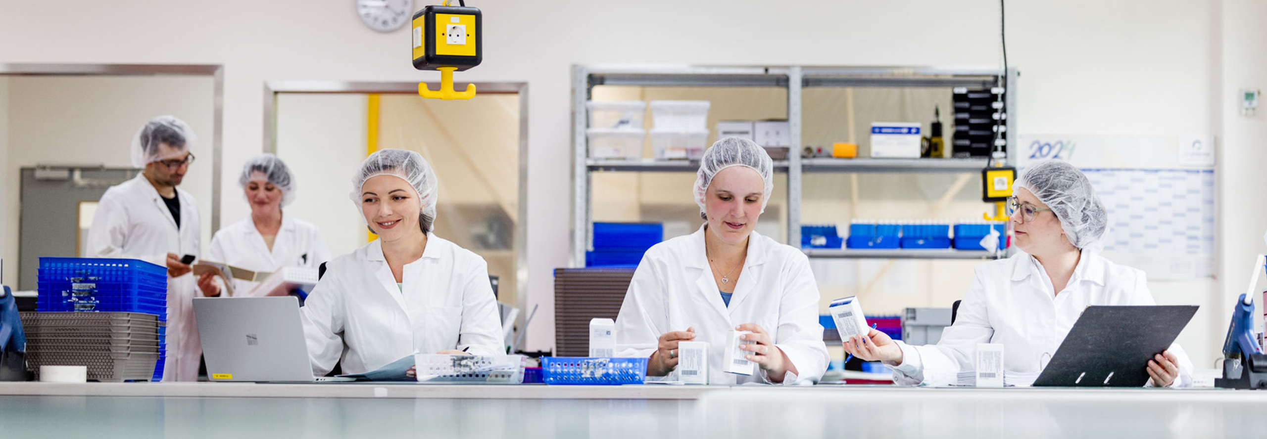 Modern laboratory environment with five professionals, four women and one man, all wearing protective clothing (white lab coat and hairnet), sitting and standing around a laboratory bench. The people in the foreground are checking the labels on pharmaceutical packaging.