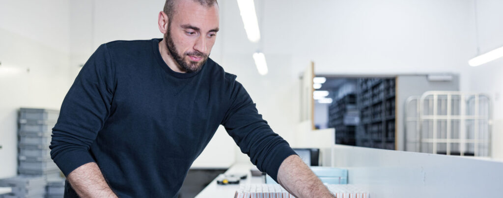 A man is working at a workstation in a warehouse-type environment. He is bending over a table on which paraphernalia like pharmaceutical packaging can be seen.