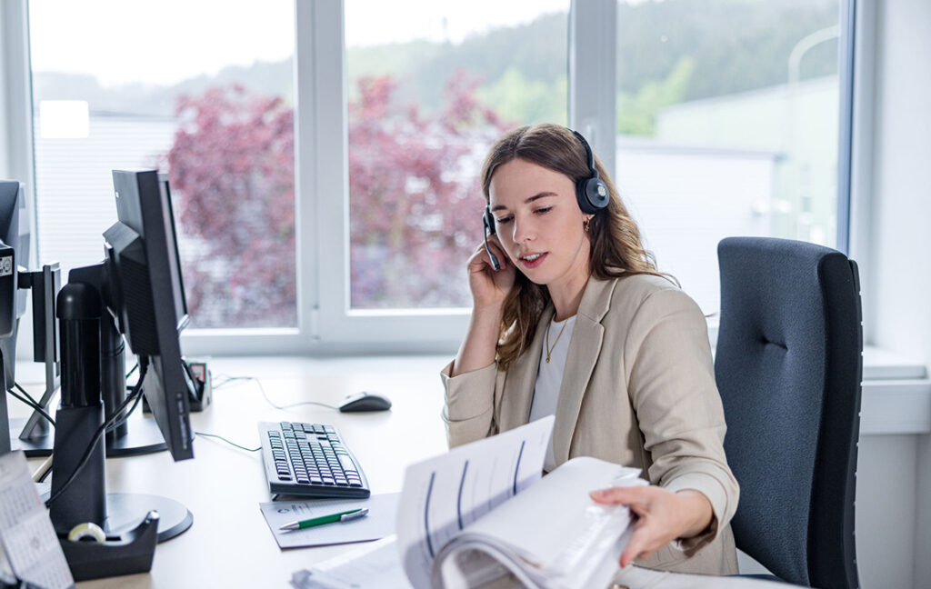 Eine Frau im beigen Blazer arbeitet vor einem Computer in einer Büroumgebung. Sie trägt ein Headset und blättert durch einen Ordner. Der Raum hat große Fenster und bietet einen Blick auf grüne Außenbereiche.