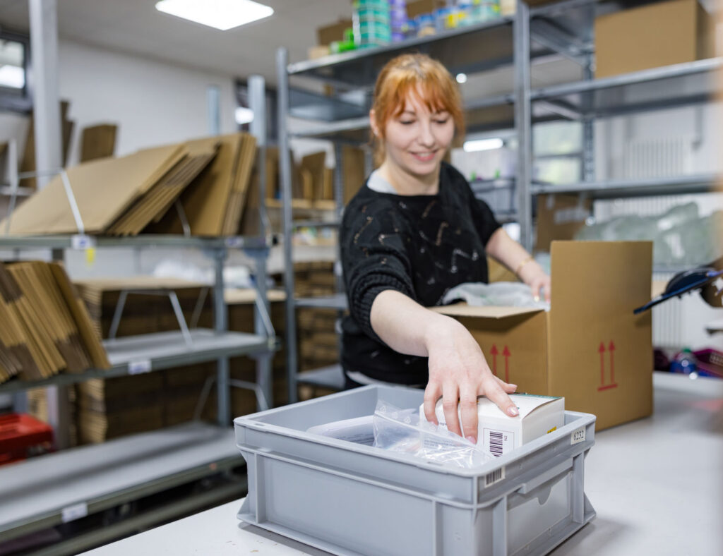 Woman with red hair smiles as she packs medicinal products in a distribution center.