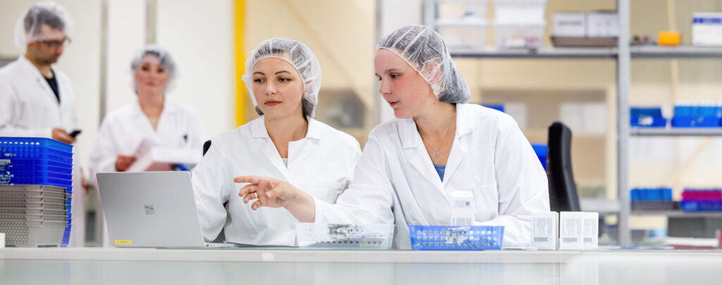 In a laboratory environment, two women can be seen in the foreground wearing white lab coats and hairnets. The woman on the left looks at a laptop screen, while the woman on the right points to it. There are two other people visible in the background, a man and a woman also wearing protective clothing. They are standing and talking to one another.