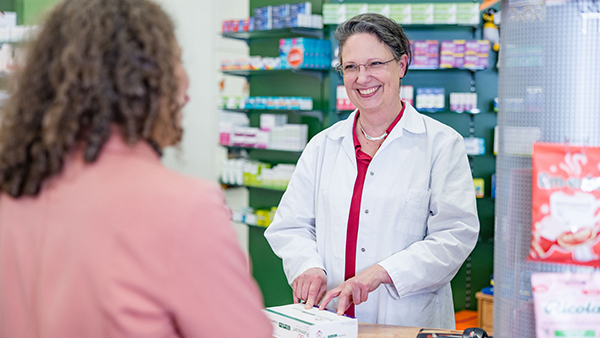 Frau in einem weißen Kittel steht hinter einem Apothekentresen und zeigt lächelnd mit ihren Händen auf eine Medikamentenverpackung. Die Kundin ist nur von hinten zu sehen. Im Hintergrund sind Medikamentenregale zu sehen.