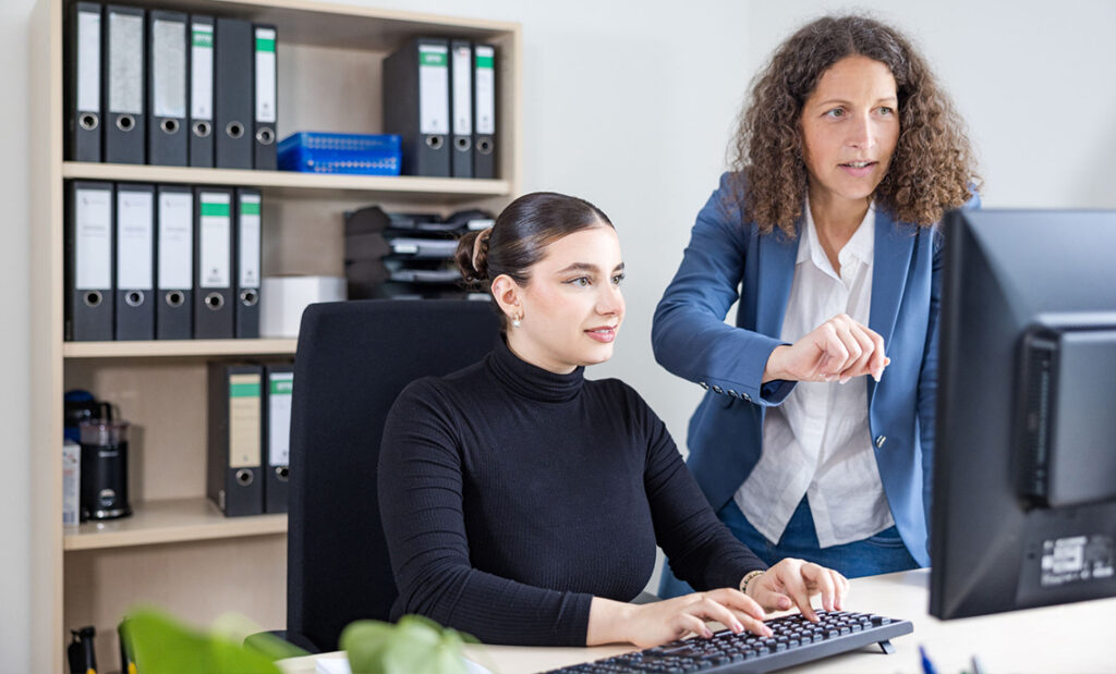 Zwei Frauen befinden sich gemeinsam in einem Büroraum. Die Frau links sitzt an einem Schreibtisch vor einem Computerbildschirm und bedient mit ihren Händen die Tastatur. Die Frau rechts steht neben ihr und zeigt auf den Bildschirm.