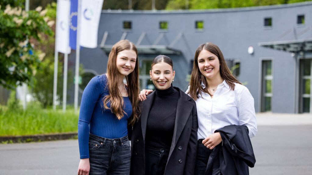 Drei junge Frauen stehen lächelnd vor dem Firmengebäude von CC Pharma und schauen in die Kamera.