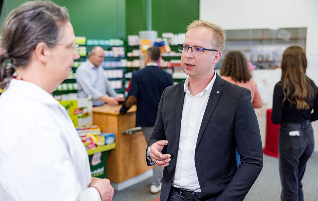 Gesprächssituation in einer Apotheke. Im Vordergrund sind eine Frau und ein Mann, die sich miteinander unterhalten. Die Frau trägt einen weißen Kittel, während der Mann im Anzug ist an dessen Revers eine CC Pharma Stecknadel zu sehen ist. Im Hintergrund sind vier weitere Personen zu sehen. Ein Mann als Mitarbeiter der Apotheke hinter der Theke, ein Mann und zwei Frauen als Kunden und Kundinnen der Apotheke auf der anderen Seite der Theke.