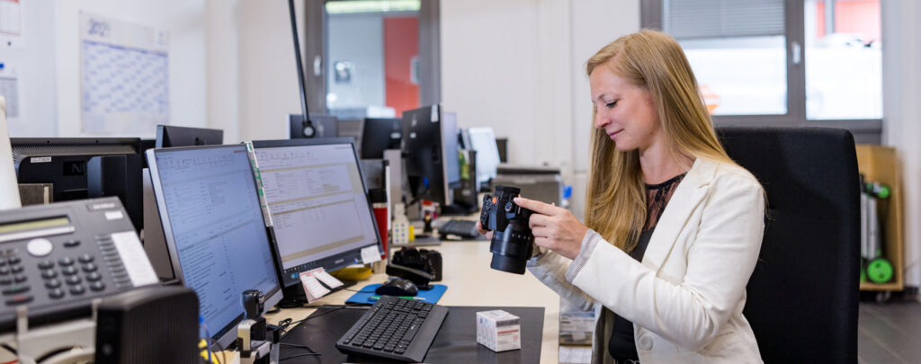 Woman in an office smiles at her camera which she is using to photograph a pack of medication. She sits in front of several monitors at a desk on which several office tools are visible.