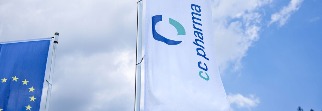 Two flags flutter against a blue sky with light clouds: one flag in white with the CC Pharma logo and part of the European Union flag.