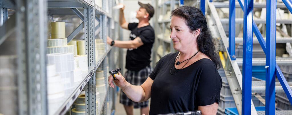 Two people, a man and a woman, stand in front of shelves in a warehouse environment, whereby the woman in the foreground is using a hand scanner. The man in the background is holding products from the shelf in his hand.