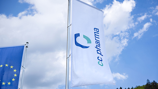 Zwei Flaggen wehen vor einem blauen Himmel mit leichten Wolken: eine Flagge in weiß mit dem Logo von CC Pharma und angeschnitten die Flagge der Europäischen Union.