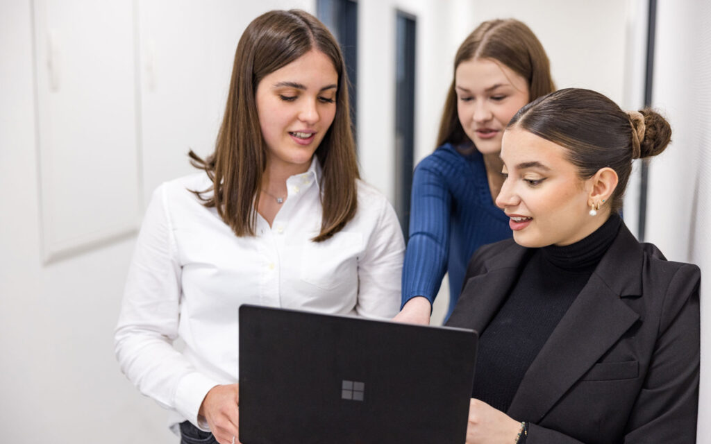 Drei junge Frauen stehen in einem Flur und schauen gemeinsam auf einen Laptop.