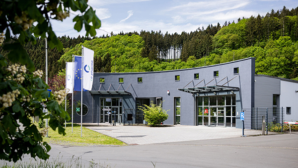 Ansicht des Firmengebäudes von CC Pharma, umgeben von grünen Bäumen und unter blauem Himmel mit wenig Wolken. Vor dem Gebäude wehen drei Flaggen, darunter eine Flagge mit dem Logo von CC Pharma, eine EU-Flagge und eine weitere Flagge, die nicht eindeutig zu erkennen ist.