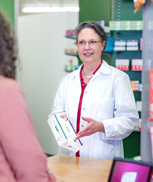 Mitarbeiterin einer Apotheke in weißem Apothekerkittel steht an einem Apothekentresen und überreicht lächelnd einer Kundin eine Packung Medikamente. Im Hintergrund sind Apothekenregale mit verschiedenen Produkten sichtbar.