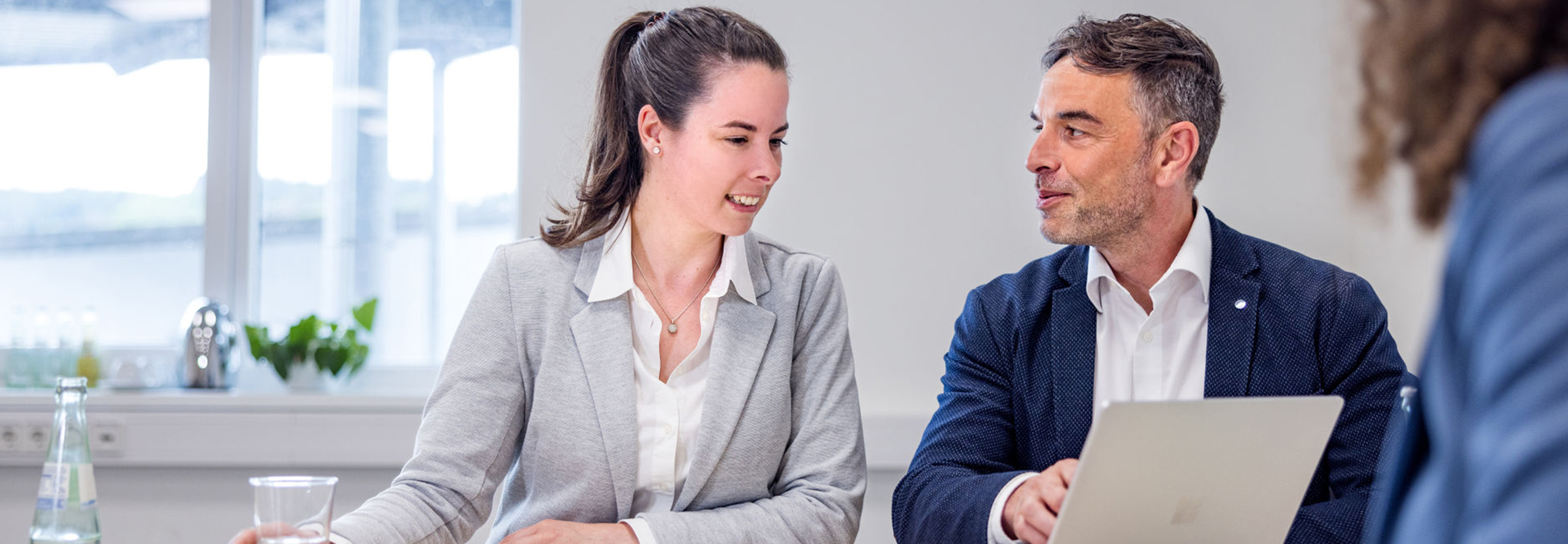 Besprechungssituation in einem hellen Büro, bei der ein Mann und eine junge Frau in Business-Kleidung, lächelnd an einem Tisch sitzen. Der Mann zeigt auf einen Laptopbildschirm, während die Frau zuschaut. Am rechten äußeren Rand des Bildes ist eine weitere Person von hinten zu erkennen.