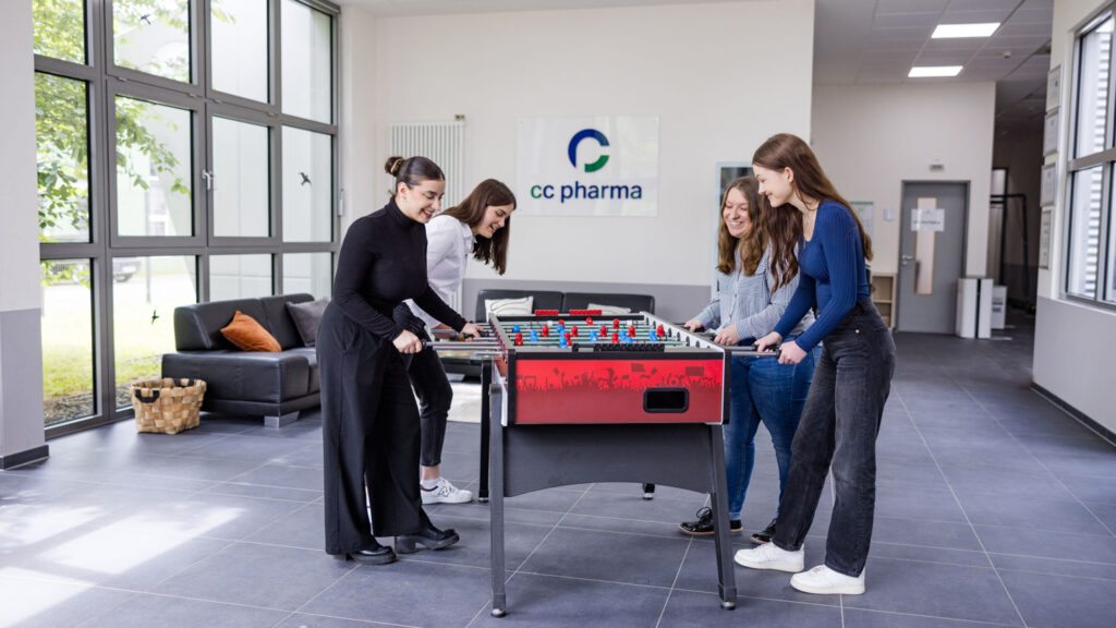 Vier junge Frauen spielen Tischfußball in einem Aufenthaltsraum und zeigen fröhliche Gesichtsausdrücke. Im Hintergrund ist das Firmenlogo von CC Pharma an der Wand sichtbar.