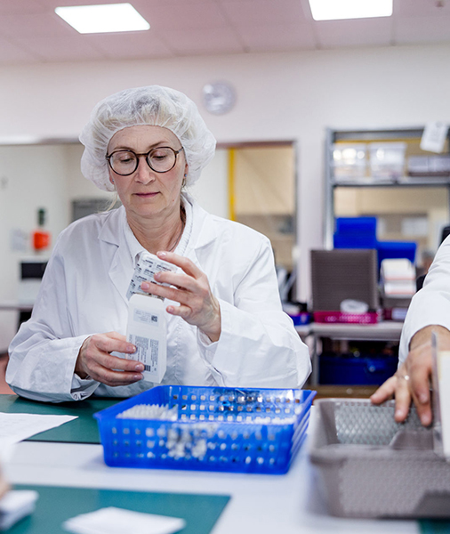 A lab worker in a white coat and wearing a hairnet looks intently at the medicines in her hand. In the foreground there are other medicines to be packaged.