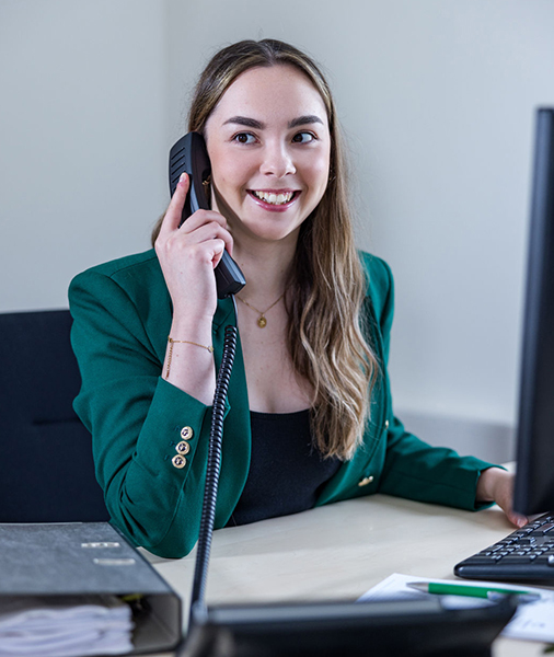 A woman in a green jacket is smiling as she sits in an office-like environment and uses the phone. She is holding a phone in her left hand while her right hand is resting on the computer keyboard.